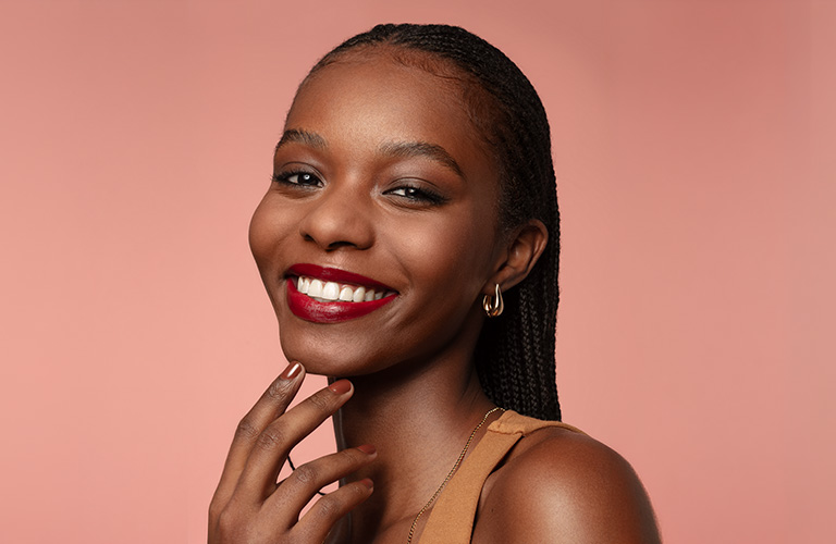 Foto de una mujer en primer plano y fondo rosa. Mano junto al rostro con uñas marrones, y cabello trenzado, top color camel y collar fino.